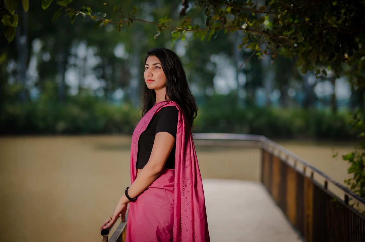 Young woman standing on a bridge wearing a pink saree, looking contemplative.