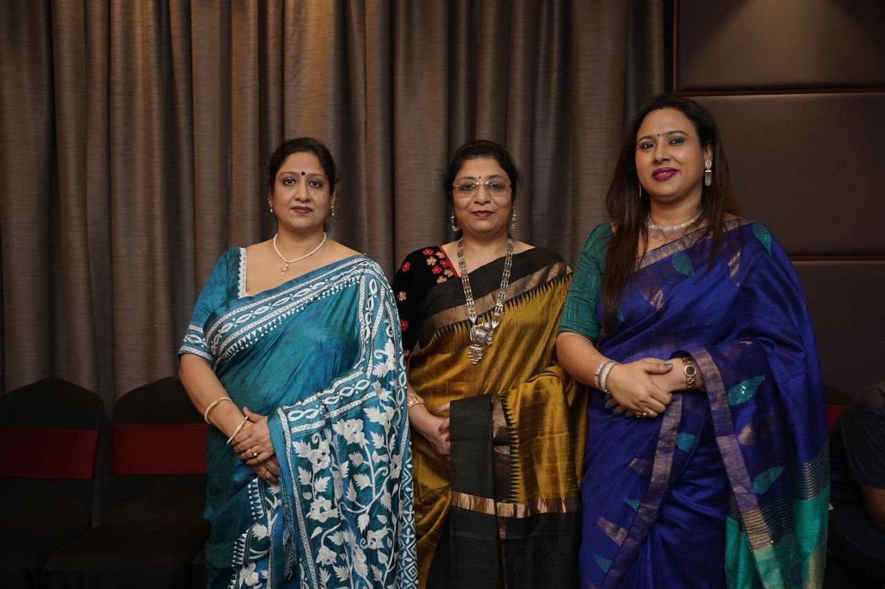 Three Indian women in colorful sarees pose gracefully indoors, showcasing traditional fashion.