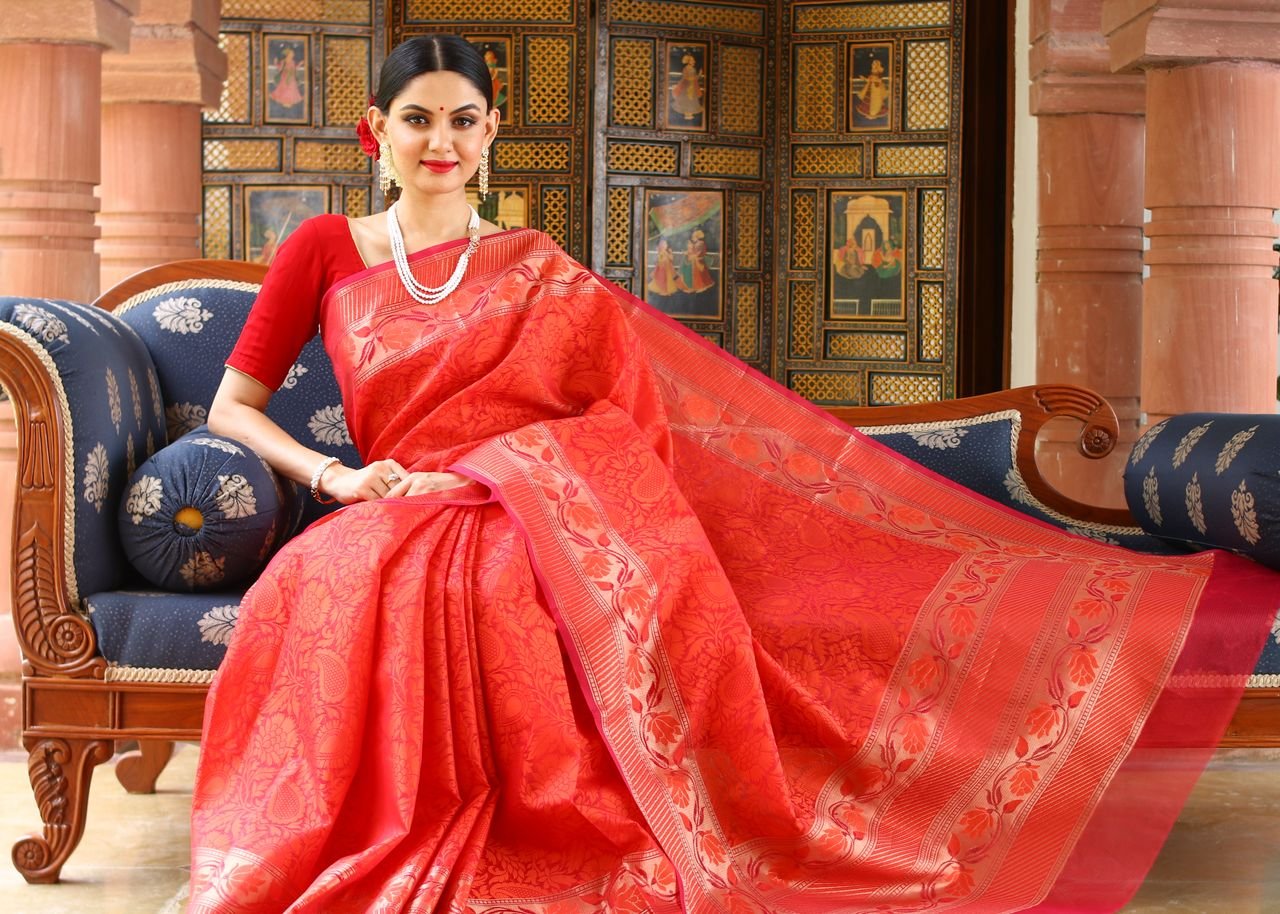 Woman in red saree poses elegantly indoors, highlighting traditional fashion and culture.